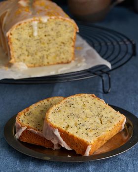 Lemon bread coated with sugar sweet icing and sprinkled with lemon peel. Slice of cake with citrus, poppy, traditional american cuisine. Dark blue table, vertical