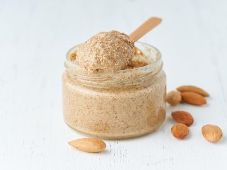 Almond butter, raw food paste made from grinding almonds into nut butter, crunchy and stir, white wooden table, glass jar, side view, close up