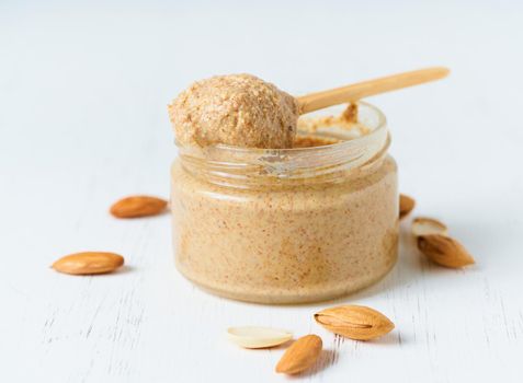 Almond butter, raw food paste made from grinding almonds into nut butter, crunchy and stir, white wooden table, glass jar, side view, close up