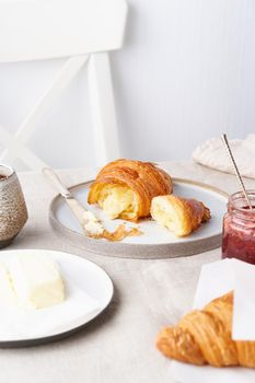 Coffee with croissant. Traditional french breakfast. Bright sunny morning, unhurried breakfast with fresh pastries, butter and jam. White wall, white chair, light tablecloth. Side view, vertical
