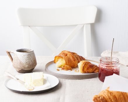 Coffee with a croissant. Bright sunny morning, unhurried breakfast with fresh pastries, butter and jam. Traditional french breakfast. White wall, white chair, light tablecloth. Side view