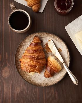 Morning breakfast with croissantand slice on plate, cup of coffee, jam and butter. Dark brown table , morning routine, slow life. Top view, vertical