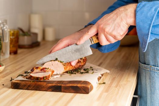 Unrecognizable man cuting with large knife baked pork tenderloin on cutting board on itchen table at home, slicing thin slices of meat with sharp blade