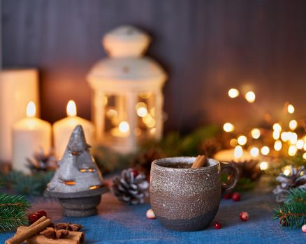 Cup of hot drink on christmas background. Cozy evening, mug of mulled wine, xmas decorations, candles and lights garlands. Dark blue backdrop with glitter and copy space