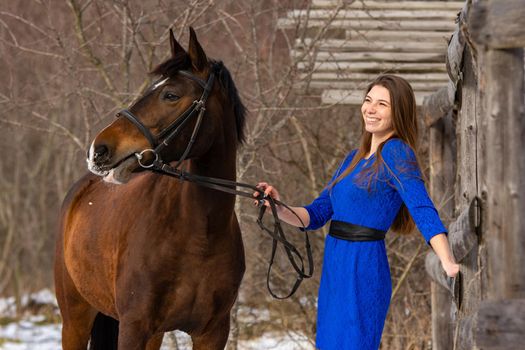 A happy girl holds a horse by the bridle and smiles, against the backdrop of a winter forest a
