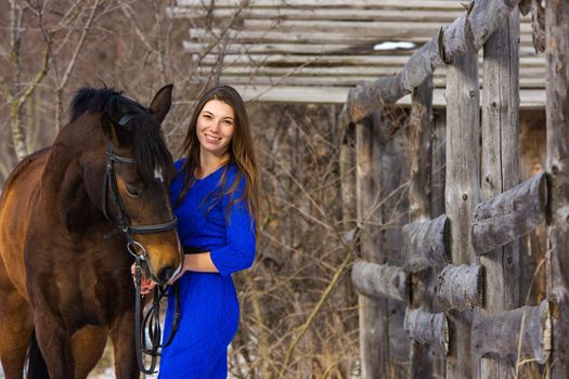 A beautiful young girl in a blue dress hugs a horse against the background of an old fence and a winter forest a