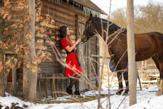 A girl in a red dress stands near an old wooden house, a horse stands next to her