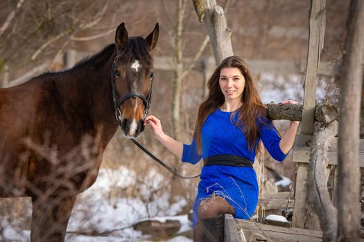 A beautiful girl is sitting on a log against the background of wooden ruins, a horse is standing nearby a
