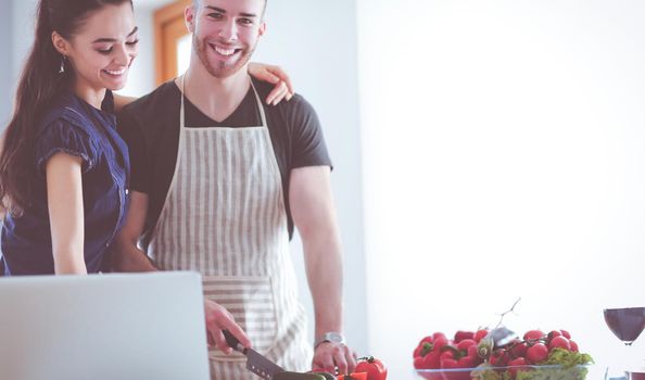 Young man cutting vegetables and woman standing with laptop in the kitchen.