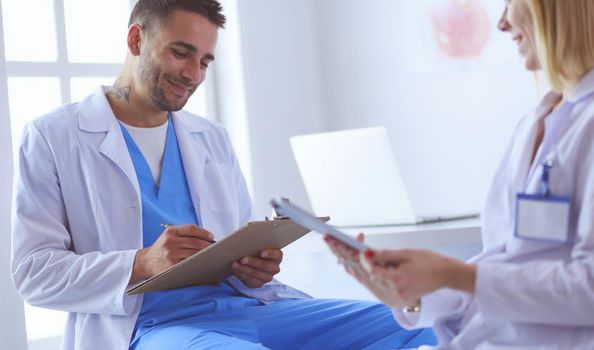 Handsome doctor is talking with young female doctor and making notes while sitting in his office