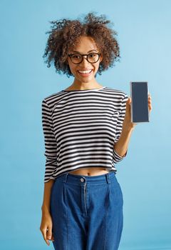 Joyful multiracial female person looking at camera and smiling while holding smartphone. Isolated on blue background