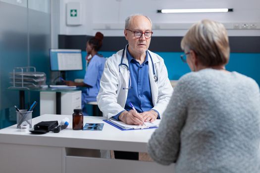 Physician signing checkup papers to give prescription treatment to ill woman. Specialist writing diagnosis on clipboard files, after doing medical consultation at checkup appointment.