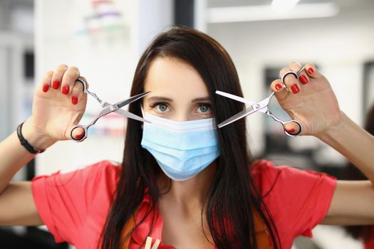 Portrait of young qualified master holding pair of scissors for work and posing in face mask. Beauty industry work during pandemic. Beauty studio concept