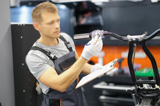 Portrait of garage service worker check wheel on motorbike, hold clipboard with info. Service center with qualified mechanic. Pit stop, repairing concept