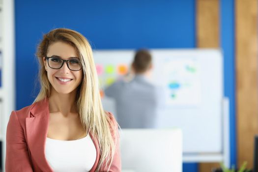 Portrait of smiling businesswoman posing in office with in presentable pink costume, colleague on background. Business, office life, career growth concept