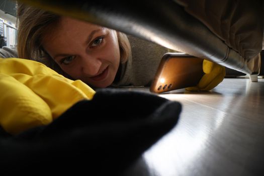 Woman cleaner looking for sock under sofa using flashlight on mobile phone. Apartment cleaning service concept