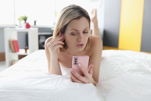 Woman lying in bed with mobile phone in her hands and inserting wireless headphones. Remote communication concept