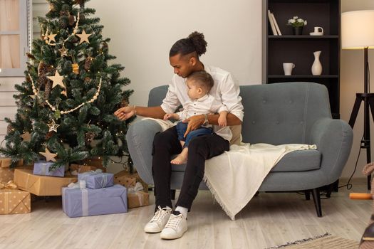 Young father and little toddler son are sitting in the living room decorated for Christmas, looking at the decorated Christmas tree