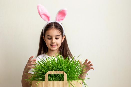 Cute little girl with pink bunny ears holding a paper bag with grass. Copy space.