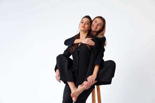 Two young business woman dressed black suit laugh and happy. One woman sitting chair and hugging other woman with barefoot legs. Positive caucasian female business people looking at camera. Isolated