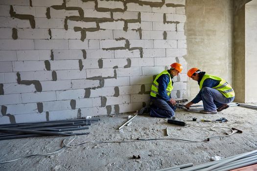 Two males professional builders in working clothes and orange safety hard hats are installing plastic pipes using modern tools in a flat of building under construction. High-rise residential house