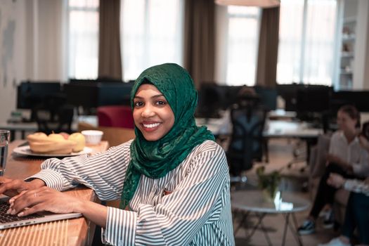 African Muslim businesswoman wearing a green hijab and working on laptop computer in relaxation area at modern open plan startup office. High-quality photo