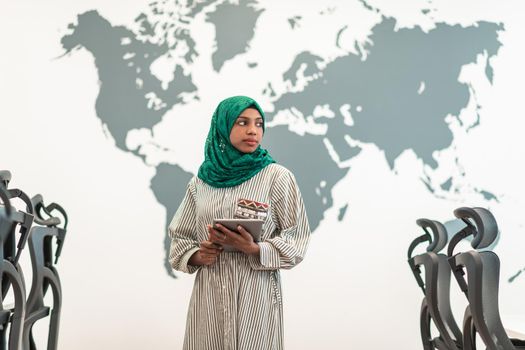 Portrait of Muslim female software developer with green hijab holding tablet computer while standing at modern open plan startup office. High-quality photo