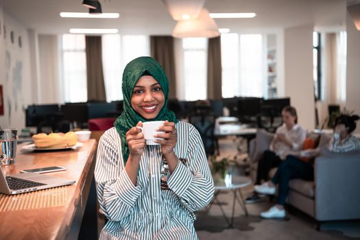 African Muslim businesswoman wearing a green hijab drinking tea while working on laptop computer in relaxation area at modern open plan startup office. High-quality photo