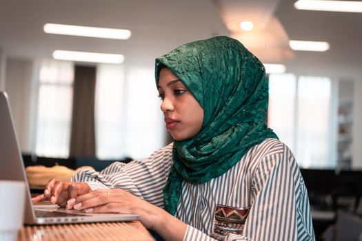 African Muslim businesswoman wearing a green hijab and working on laptop computer in relaxation area at modern open plan startup office. High-quality photo