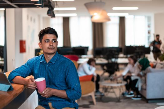 Casual businessman taking a break from the work using a laptop while drinking tea in relaxation area of modern open plan startup office. High-quality photo