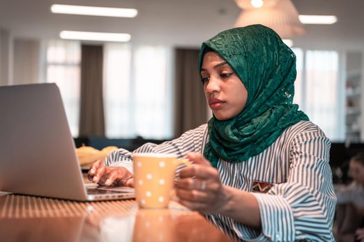 African Muslim businesswoman wearing a green hijab drinking tea while working on laptop computer in relaxation area at modern open plan startup office. High-quality photo