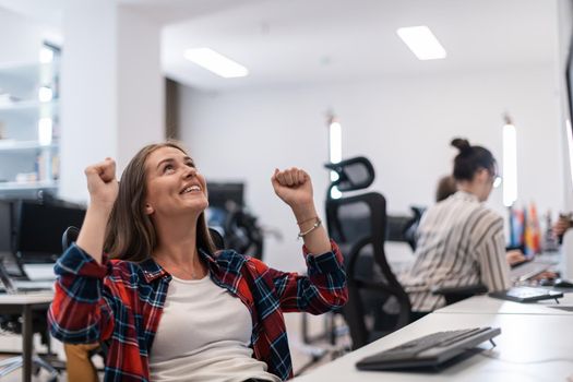 Happy casual businesswoman celebrating success while working on a desktop computer in a modern open plan startup office interior. Selective focus. High-quality photo