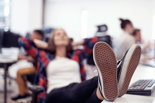 Casual businesswoman taking a break with legs on her table while working on a desktop computer in modern open plan startup office interior. Selective focus. High-quality photo