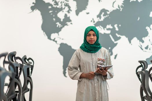 Portrait of Muslim female software developer with green hijab holding tablet computer while standing at modern open plan startup office. High-quality photo