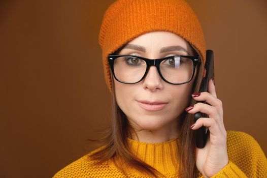 Friendly attractive caucasian woman in glasses and a knitted hat with a sweater is talking on the phone while looking at the camera. Studio portrait. Copy space. Optics and vision problems.