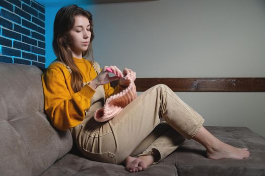 cute young caucasian woman sitting on sofa at home and crocheting wool product.