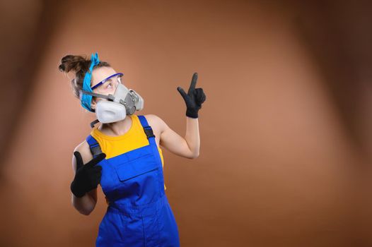 a young athletic caucasian woman, in construction overalls, a respirator, goggles and gloves, stands on a colored background. layout for mock up