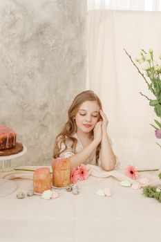 A girl with long hair in a light dress is sitting at the Easter table with cakes, spring flowers and quail eggs. Happy Easter celebration