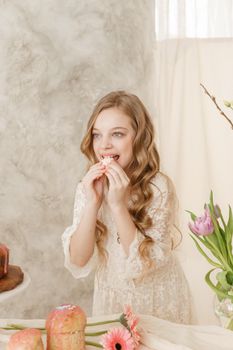 A girl with long hair in a light dress is sitting at the Easter table with cakes, spring flowers and quail eggs. Happy Easter celebration