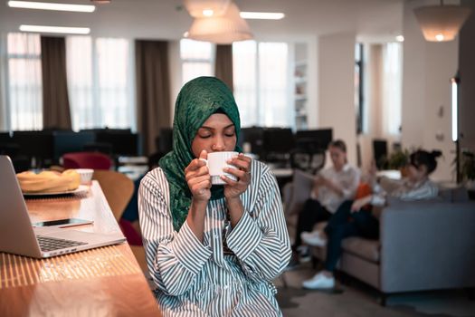 African Muslim businesswoman wearing a green hijab drinking tea while working on laptop computer in relaxation area at modern open plan startup office. High-quality photo