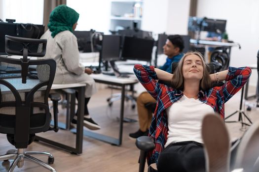 Casual businesswoman taking a break with legs on her table while working on a desktop computer in modern open plan startup office interior. Selective focus. High-quality photo