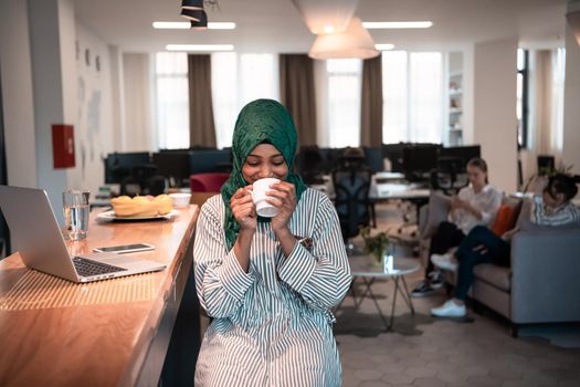 African Muslim businesswoman wearing a green hijab drinking tea while working on laptop computer in relaxation area at modern open plan startup office. High-quality photo