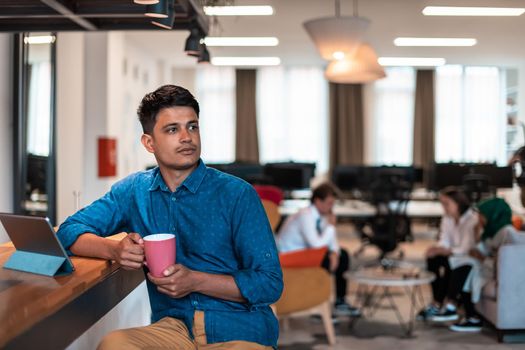 Casual businessman taking a break from the work using a laptop while drinking tea in relaxation area of modern open plan startup office. High-quality photo