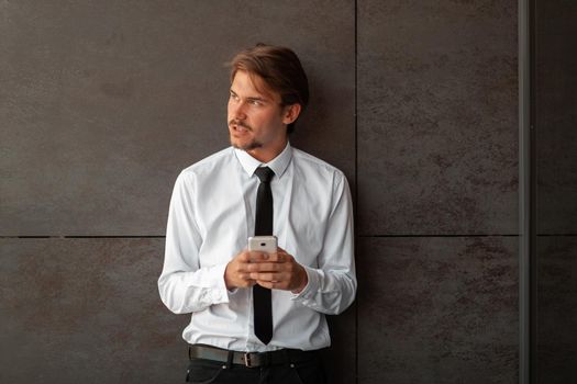 Startup businessman in a white shirt with a black tie using smartphone while standing in front of gray wall during a break from work outside. High-quality photo