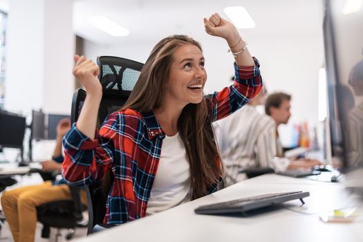 Happy casual businesswoman celebrating success while working on a desktop computer in a modern open plan startup office interior. Selective focus. High-quality photo