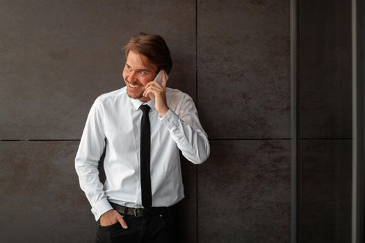 Startup businessman in a white shirt with a black tie using smartphone while standing in front of gray wall during a break from work outside. High-quality photo