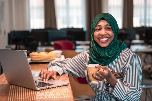 African Muslim businesswoman wearing a green hijab drinking tea while working on laptop computer in relaxation area at modern open plan startup office. High-quality photo