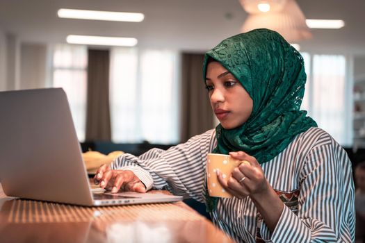 African Muslim businesswoman wearing a green hijab drinking tea while working on laptop computer in relaxation area at modern open plan startup office. High-quality photo