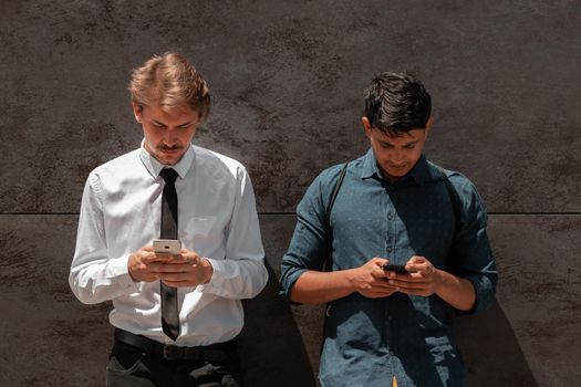 Casual multiethnic startup business men one of them is Indian using smartphone during a break from work in front of the grey wall outside. High-quality photo