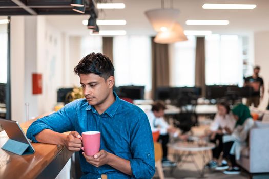 Casual businessman taking a break from the work using a laptop while drinking tea in relaxation area of modern open plan startup office. High-quality photo